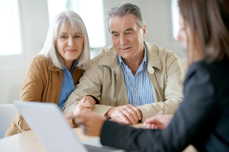 Elderly couple consulting with a professional advisor about estate planning. The couple, dressed in casual yet professional attire, is attentively reviewing documents on a laptop, indicating a detailed discussion on wills, trusts, and other estate planning services. The bright, professional setting underscores the importance of thorough estate planning for peace of mind and financial security.
